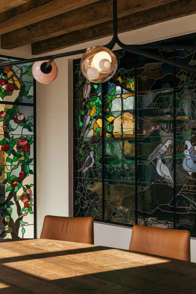 Glas-in-loodpanelen met klimmende vruchtbomen en duiven, gevat in slanke stalen kaders naast de eettafel.