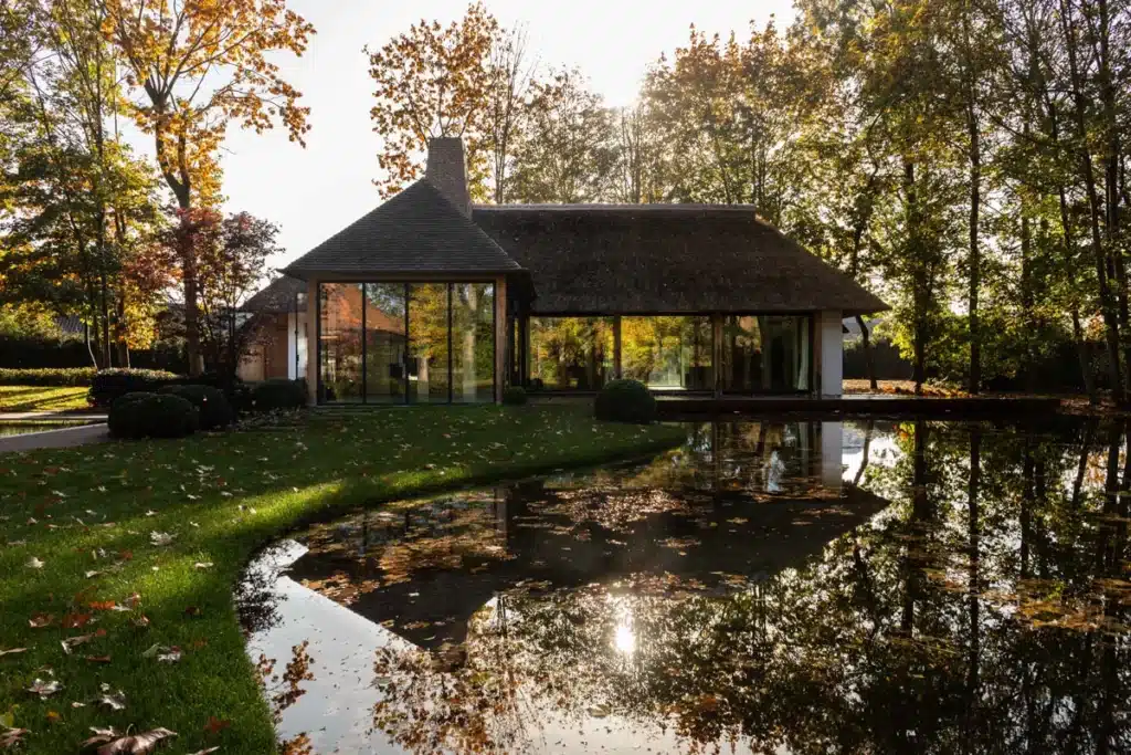 Bijgebouw met stalen glaswanden dat zich spiegelt in de vijver, tussen herfstbomen en een groen gazon.