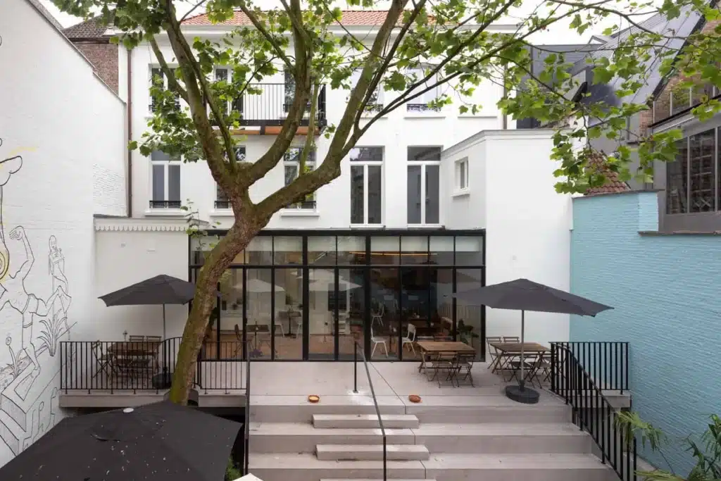 Binnentuin met boom, parasols en terras, met achteraan de stalen achtergevel en de stalen balustrades.