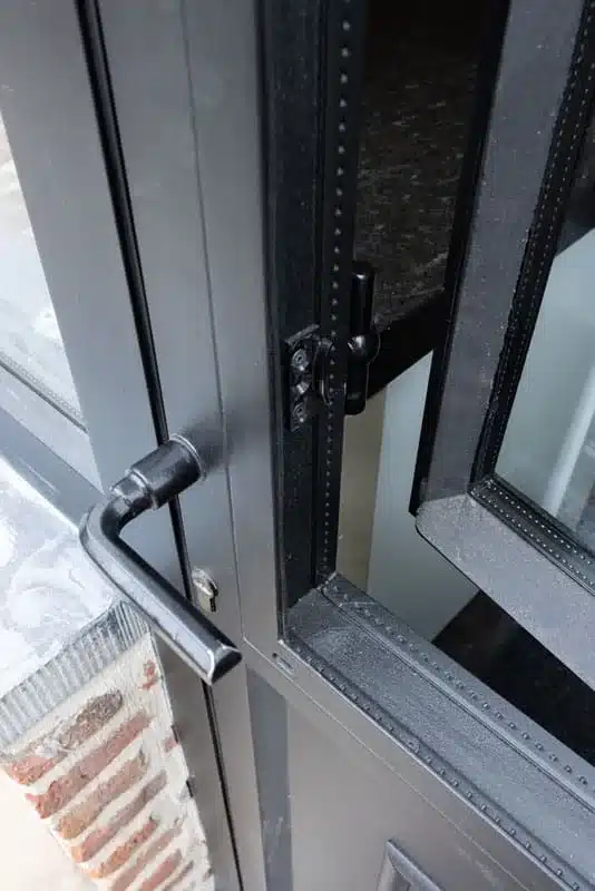 Detail van zwart gepoedercoat staal: een deurkruk, de raamsluiting en een openstaand draairaam in de deur.