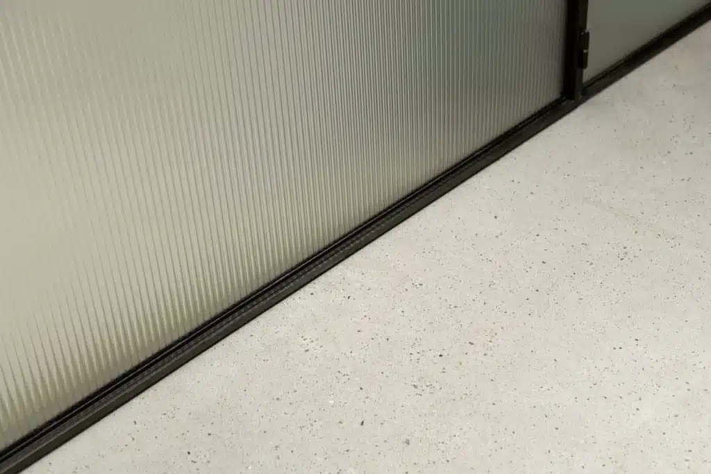 Detail van de onderkant van een stalen schuifdeur met geribd glas, met de geleiding verzonken in de vloer.