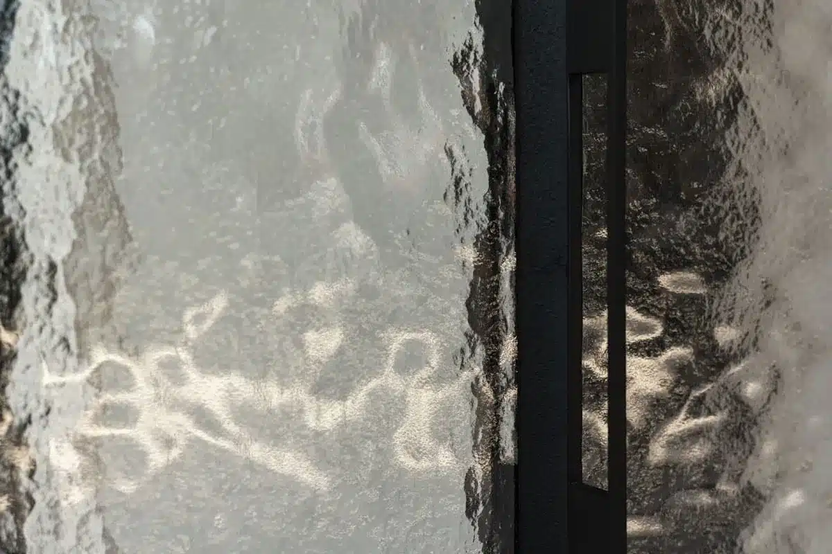 Detail van golvend structuurglas naast een zwart stalen profiel met ingewerkte greep, in het tegenlicht.