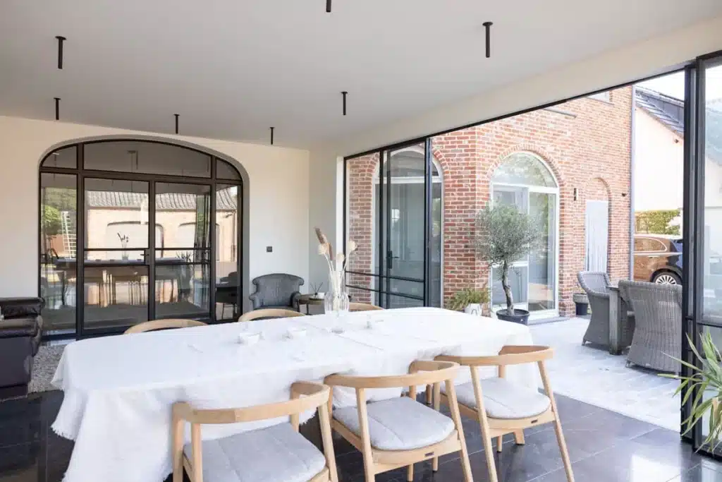 Eettafel in de tuinkamer met zicht op een stalen binnenpui met rondboog naar de woning en de open harmonicadeuren.