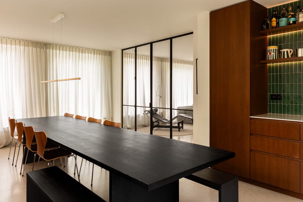 Eetkamer met zwarte tafel en houten kast, met de stalen glaswand naar de living ernaast.