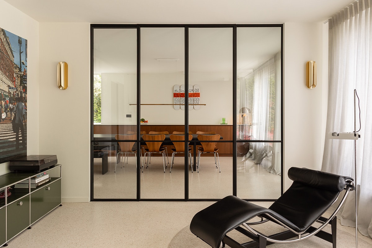 Stalen glaswand met dubbele deur tussen living en eetkamer, met een zwarte ligstoel en fijne profielen.