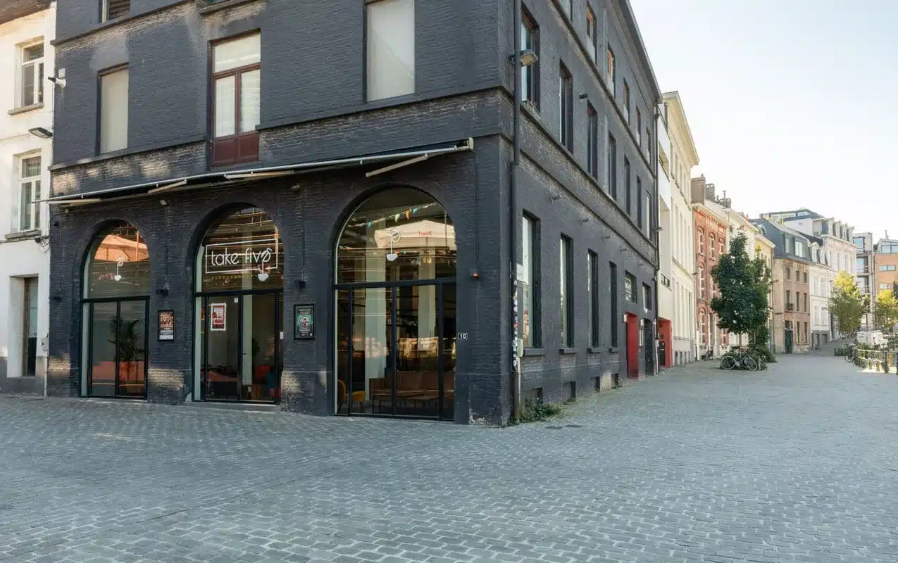 Donkere gevel van een koffiebar met drie hoge thermisch onderbroken stalen boogramen aan een kasseistraat.