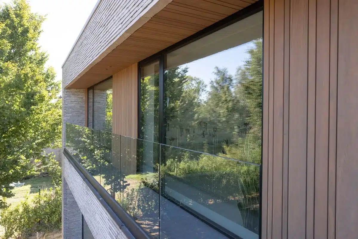 Glazen balustrade in een ingewerkt profiel langs het terras op de verdieping, tegen een houten gevel.
