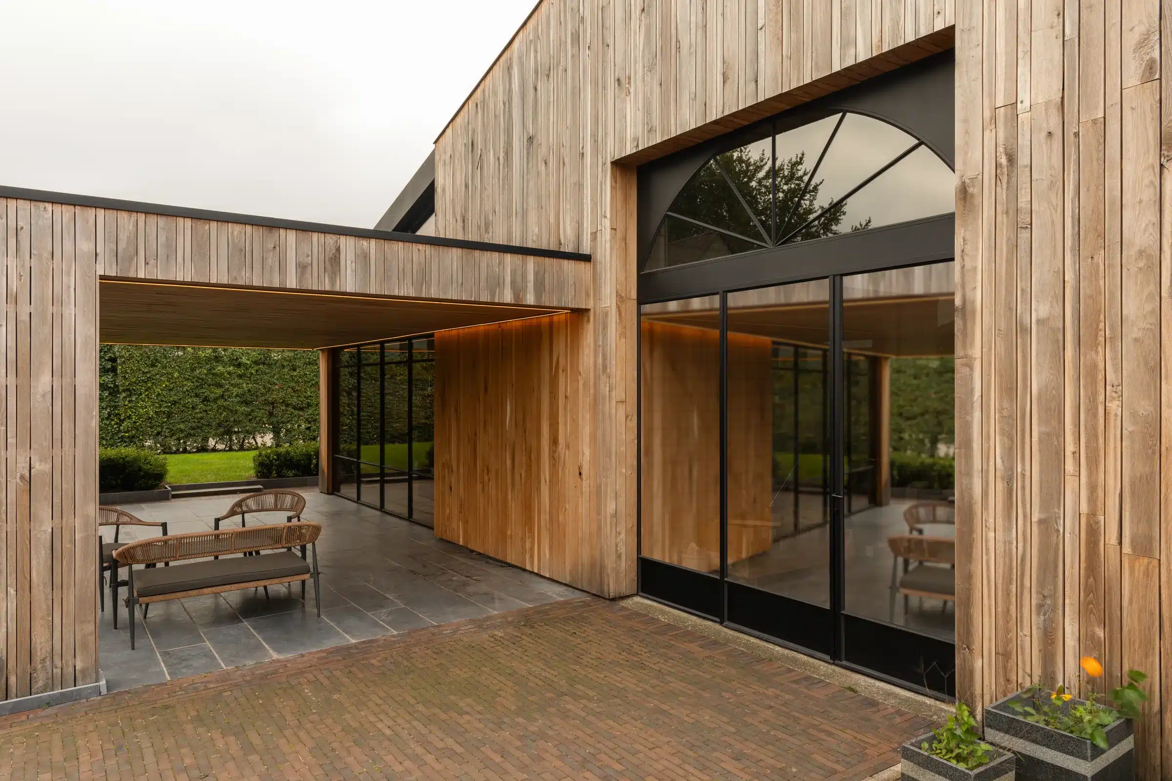 Houten gevelvolumes rond een binnenplaats, met een stalen boograam en een overdekt terras met stalen glaspui.