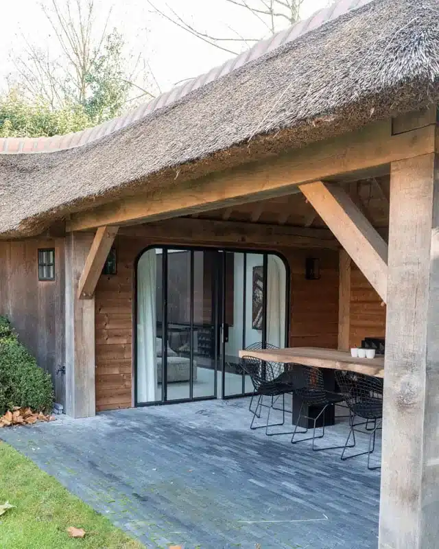 Poolhouse met rieten dak en eiken gebinte, met achter het terras de zwarte stalen raampartij en eettafel.