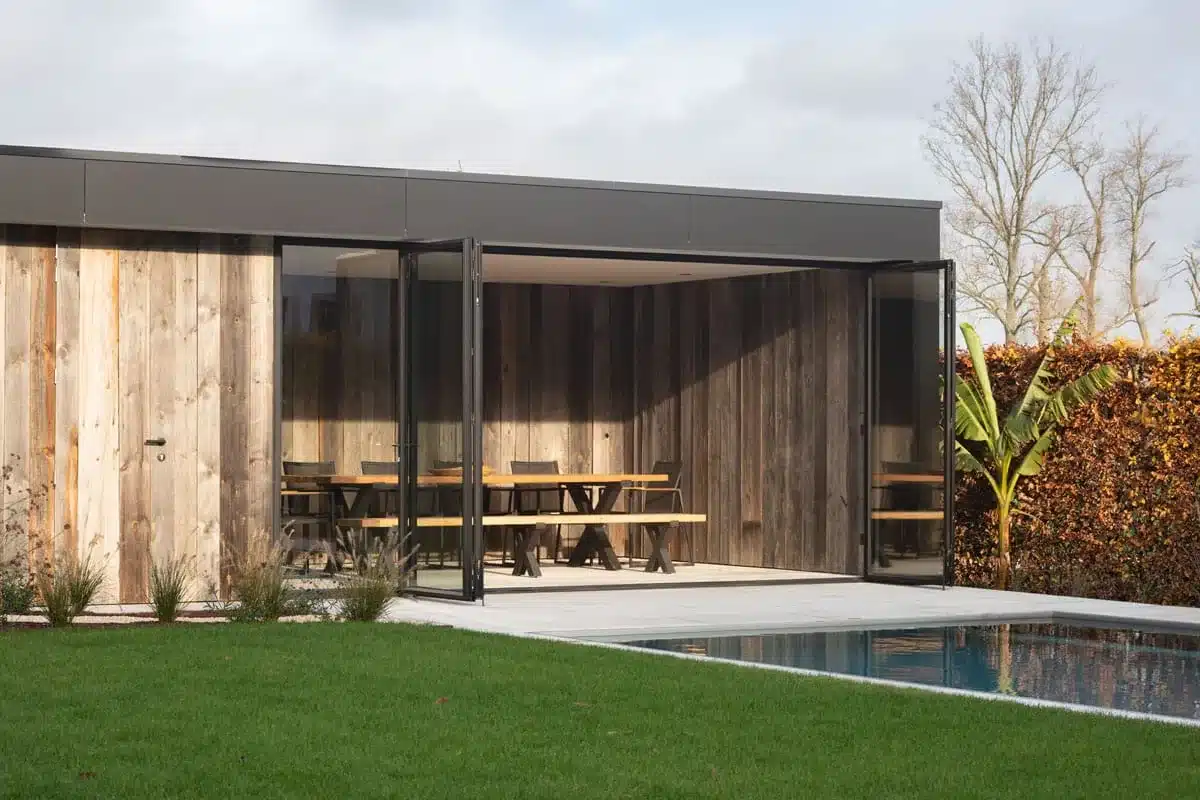 Poolhouse met opengevouwen stalen harmonicadeuren en een gevel in oud hout, aan het zwembad.