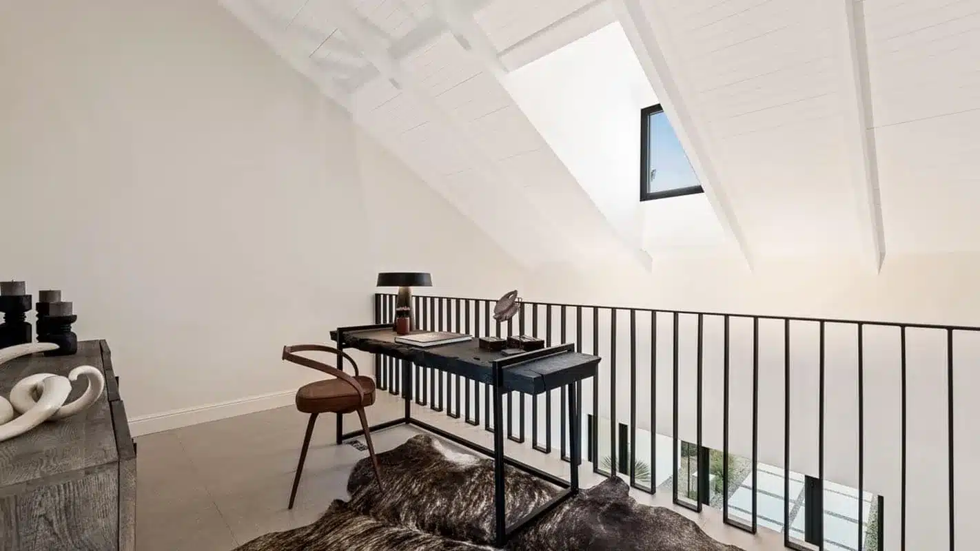 Stalen balustrade met verticale spijlen langs de vide op de verdieping, naast een bureau onder het dakraam.