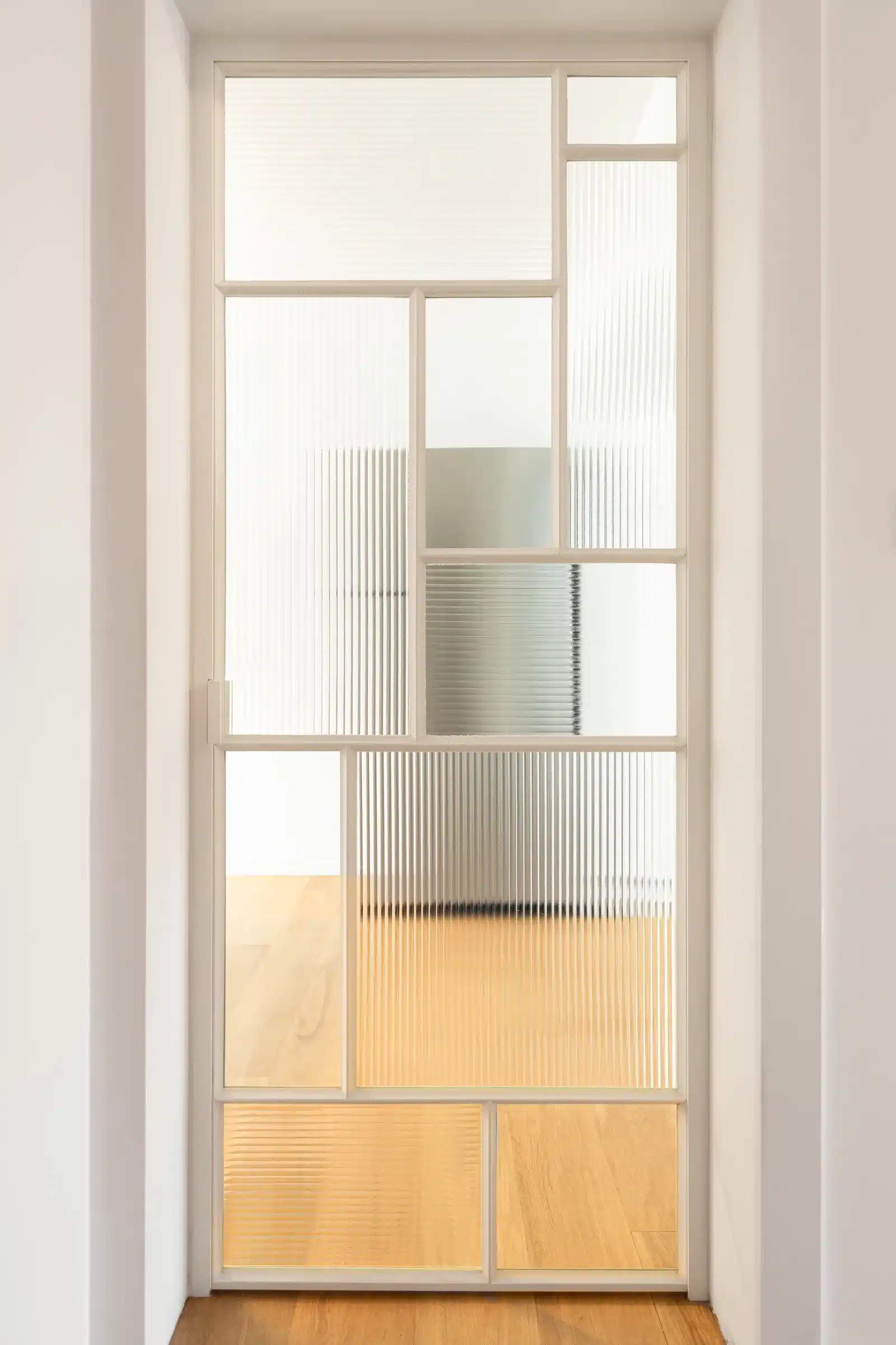 Frontale doorkijk door een deurkader naar de stalen binnendeur, met licht dat door het cannelé glas valt.