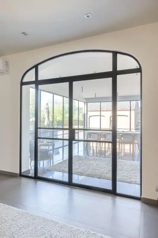 Staand zicht op de stalen binnenpui met rondboog en dubbele glazen deur, gezien vanuit de woning.