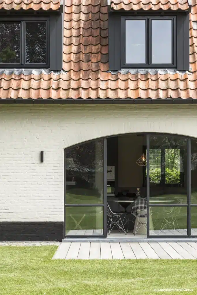 Stalen boogpui met deuren in de witgekalkte gevel van de hoeve, boven een houten terras aan het gazon.