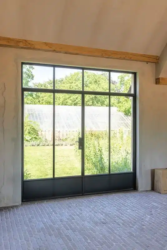 Stalen deur met zijlicht en bovenlicht onder een eiken balk, met zicht op de serre en de tuin.