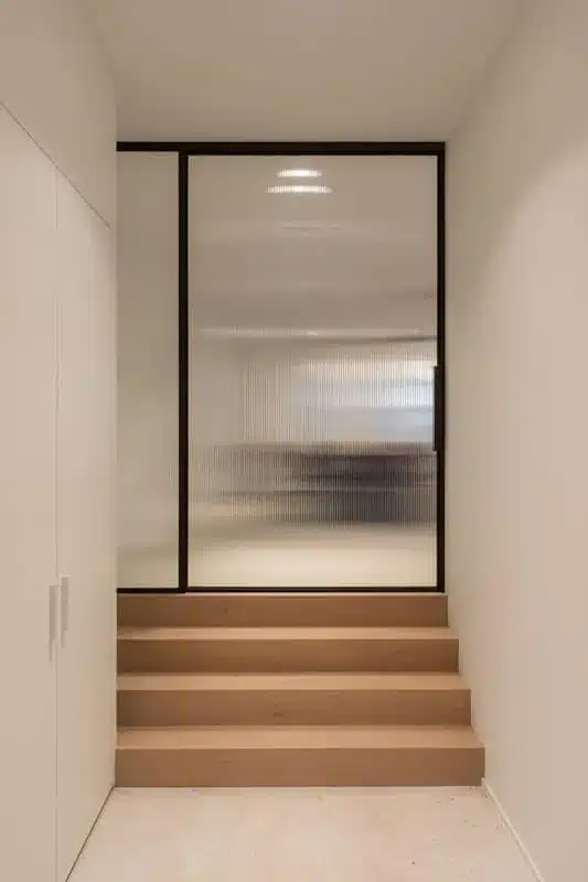 Staand zicht door een smalle gang met houten treden naar een stalen deur met geribd glas.