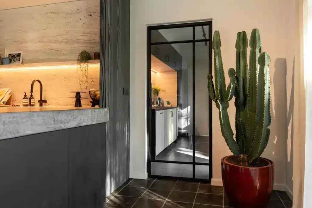 Stalen deur met glas tussen de keuken en de gang, naast een werkblad in natuursteen en een grote cactus.