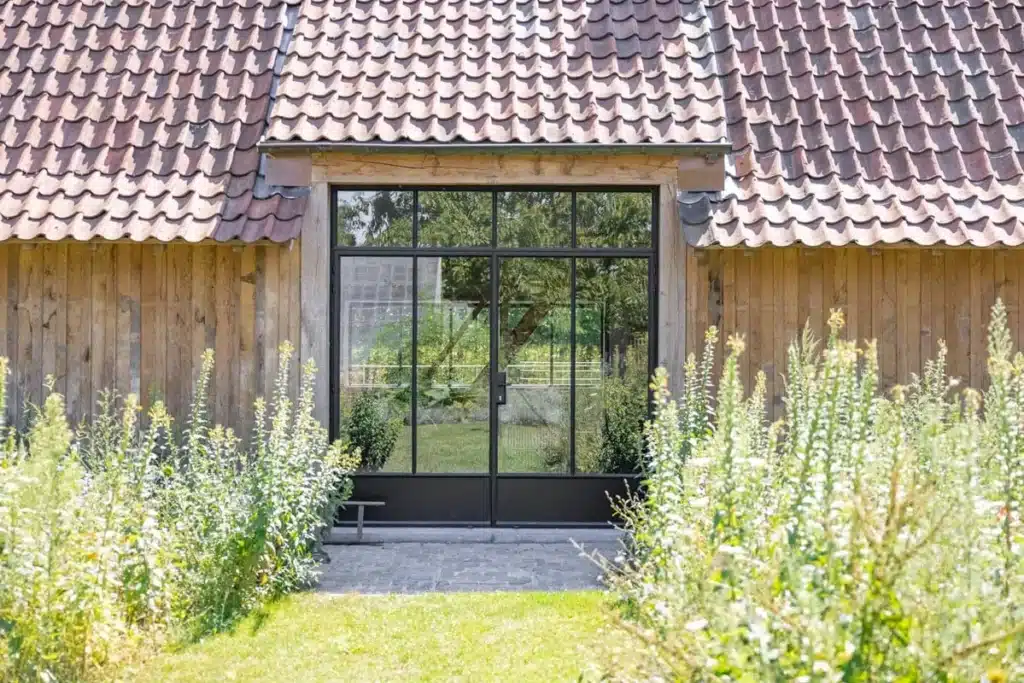 Stalen deurpartij met zijlichten in de kopgevel van het paviljoen, tussen de wilde beplanting van de tuin.