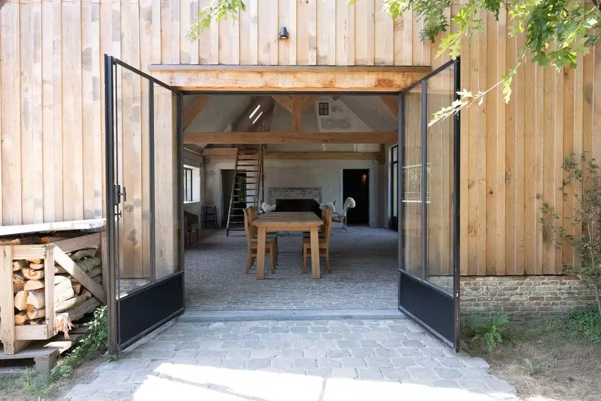 Geopende stalen dubbele deur in de houten gevel van het paviljoen, met doorzicht naar de eettafel binnen.