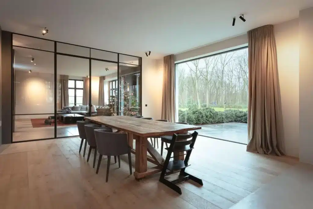Eetkamer met houten tafel naast een stalen glaswand naar de living, met groot raam op de tuin.