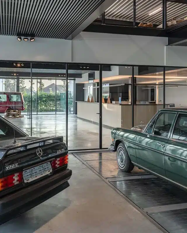 Staand zicht op de showroom met stalen glaswanden, de onthaalbalie en twee oldtimers op de voorgrond.