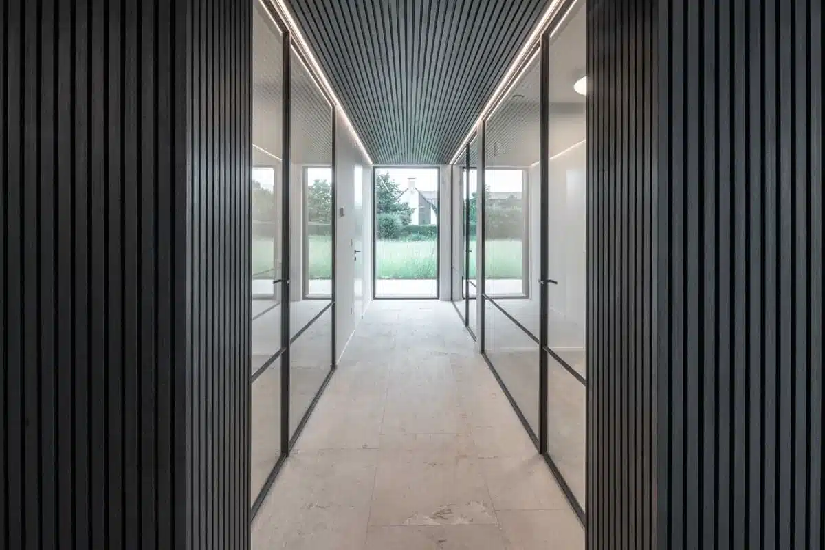 Gang tussen de kantoren met stalen glaswanden in superslanke kokerprofielen en een zwart latjesplafond.