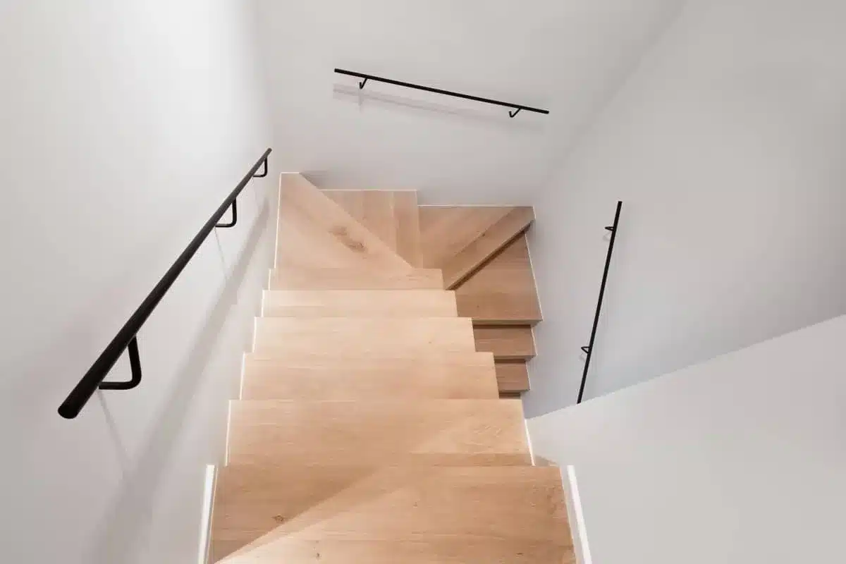 Twee zwarte stalen handgrepen zonder rozetten langs een houten trap, van bovenaf gezien in het trappenhuis.