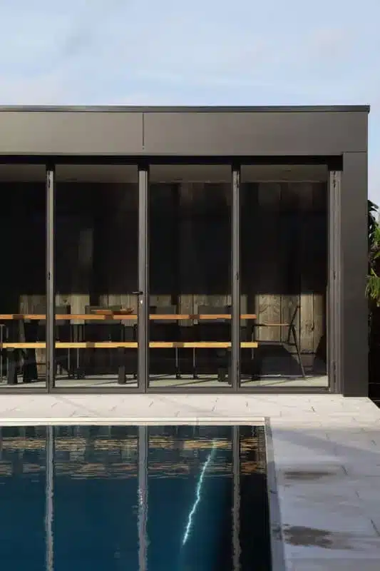 Frontaal zicht op de gesloten stalen harmonicadeuren van het poolhouse, boven de rand van het zwembad.