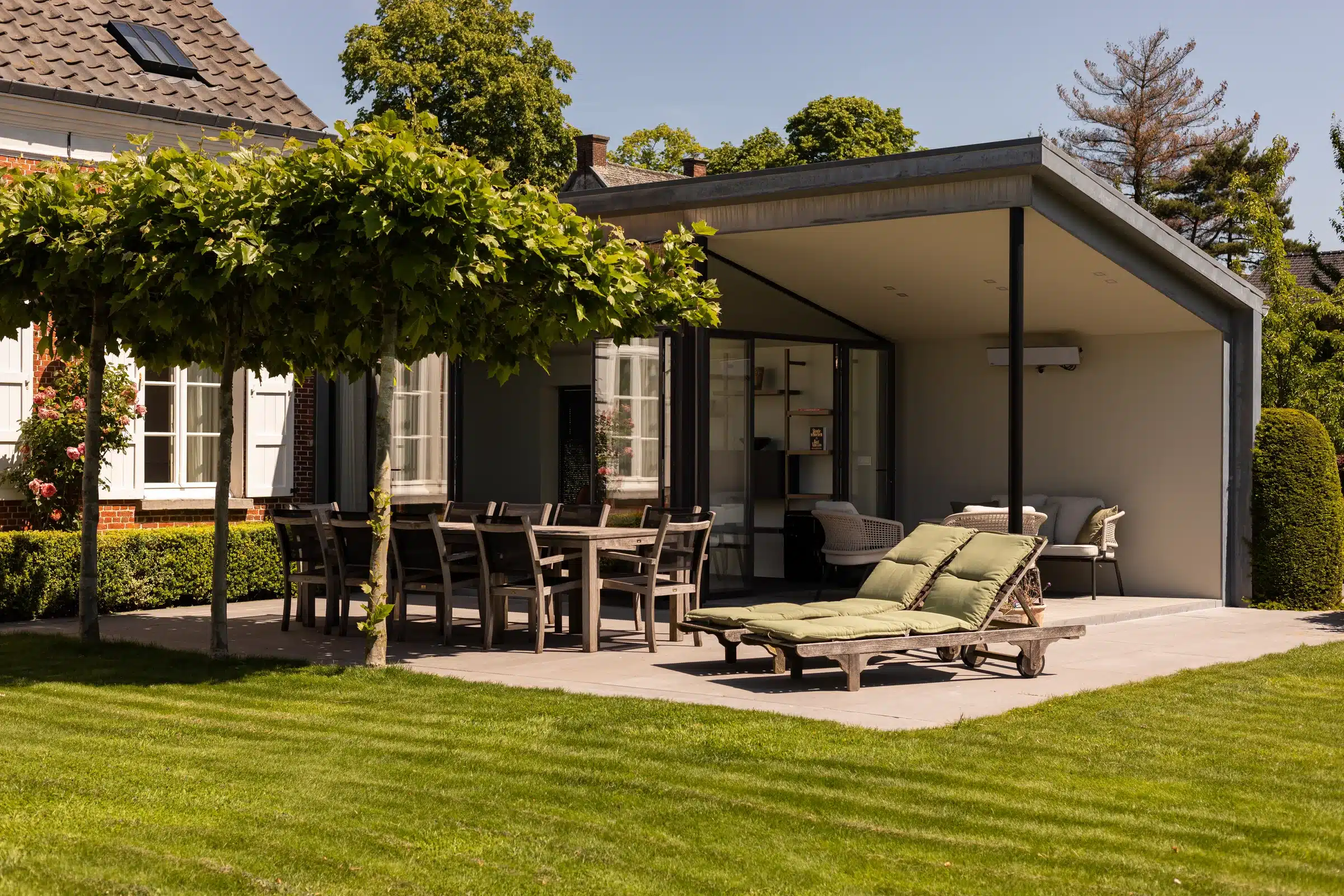 Stalen leefveranda met zwarte profielen aan de woning, met overdekt terras en zicht op de tuin.