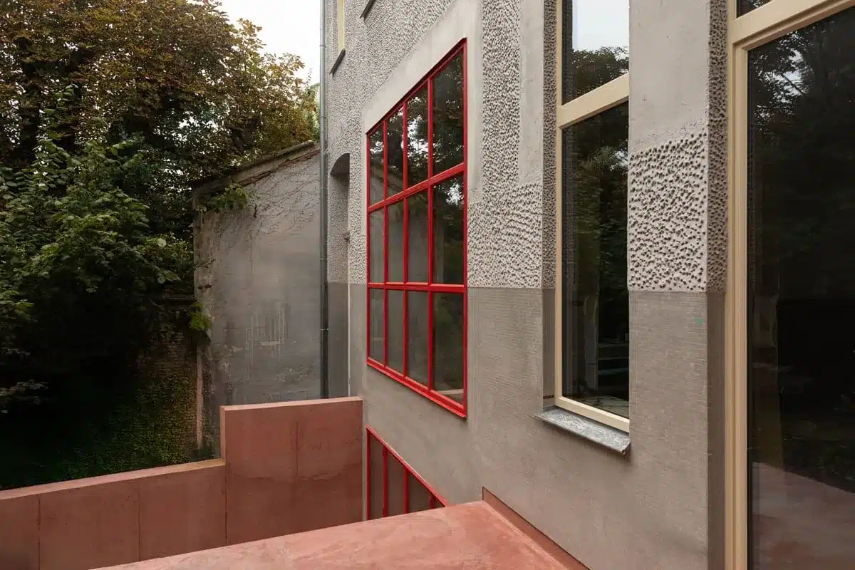 Zijzicht op de achtergevel met een rood stalen raam, de roze betonnen patio en het groen van de stadstuin erachter.