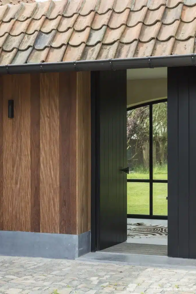 Staand zicht door een houten poort op een stalen raamdeur met rondboog, met de tuin erachter.