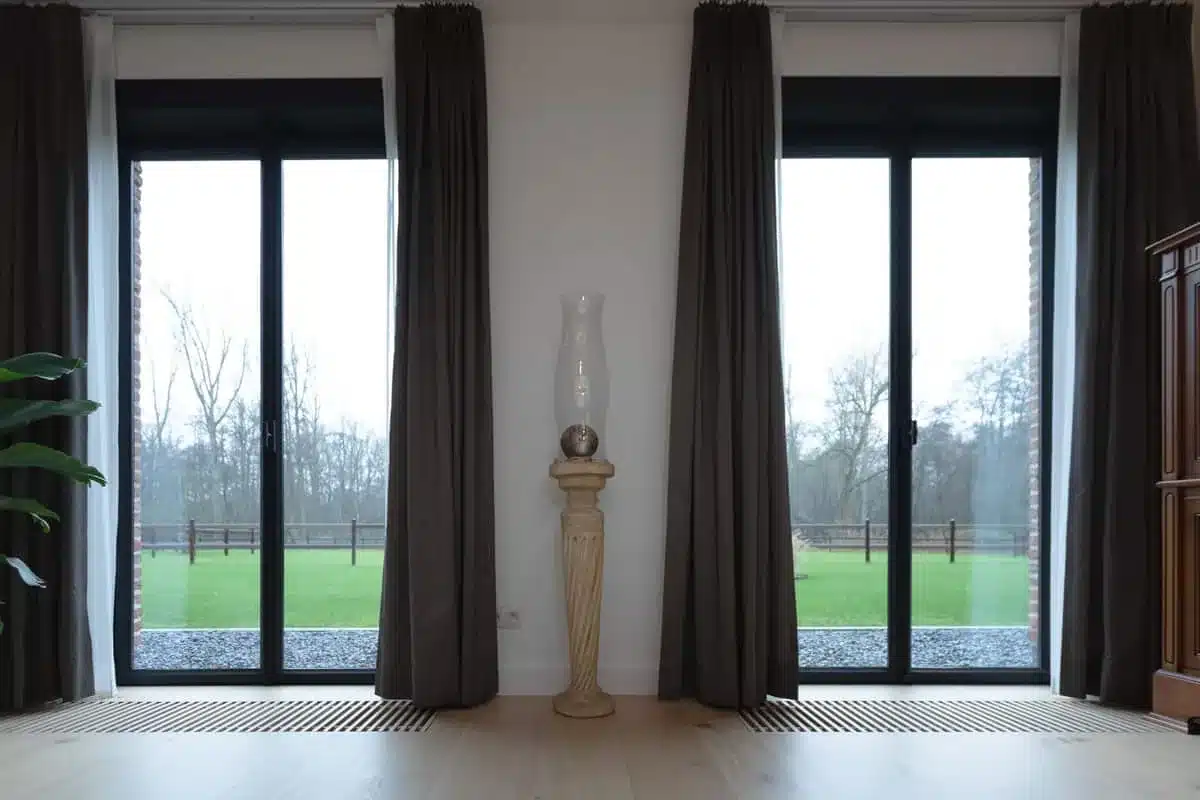 Twee hoge stalen raamdeuren in de leefruimte van de hoeve, met zicht op de omheinde weide erachter.