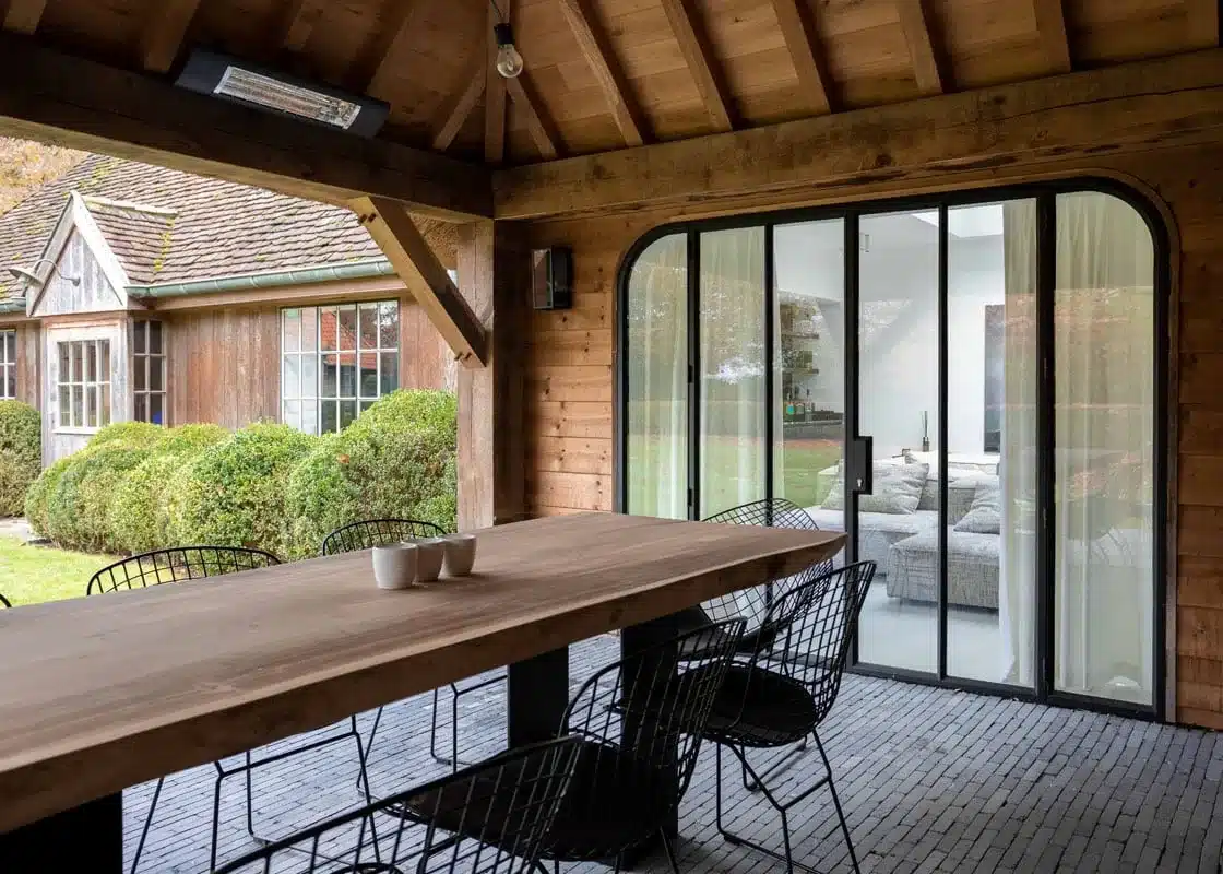 Overdekt terras onder een eiken gebinte met een brede stalen raampartij met afgeronde hoeken naar het poolhouse.
