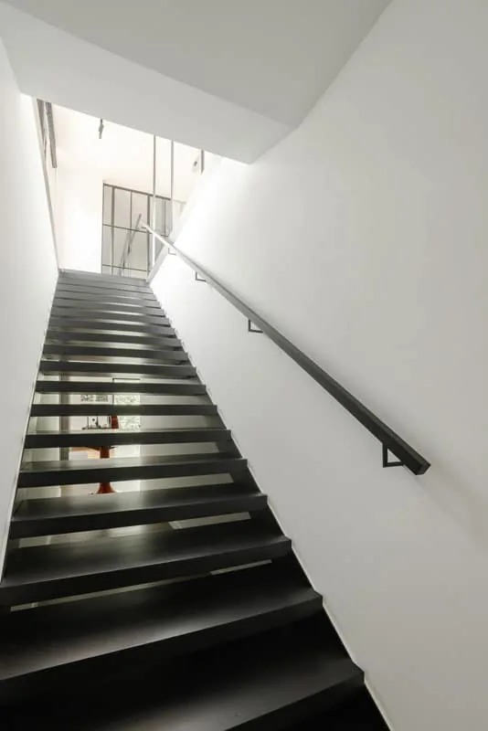 Stalen trap met zwarte treden en een platte stalen muurleuning, van onderuit gezien naar de lichte overloop.