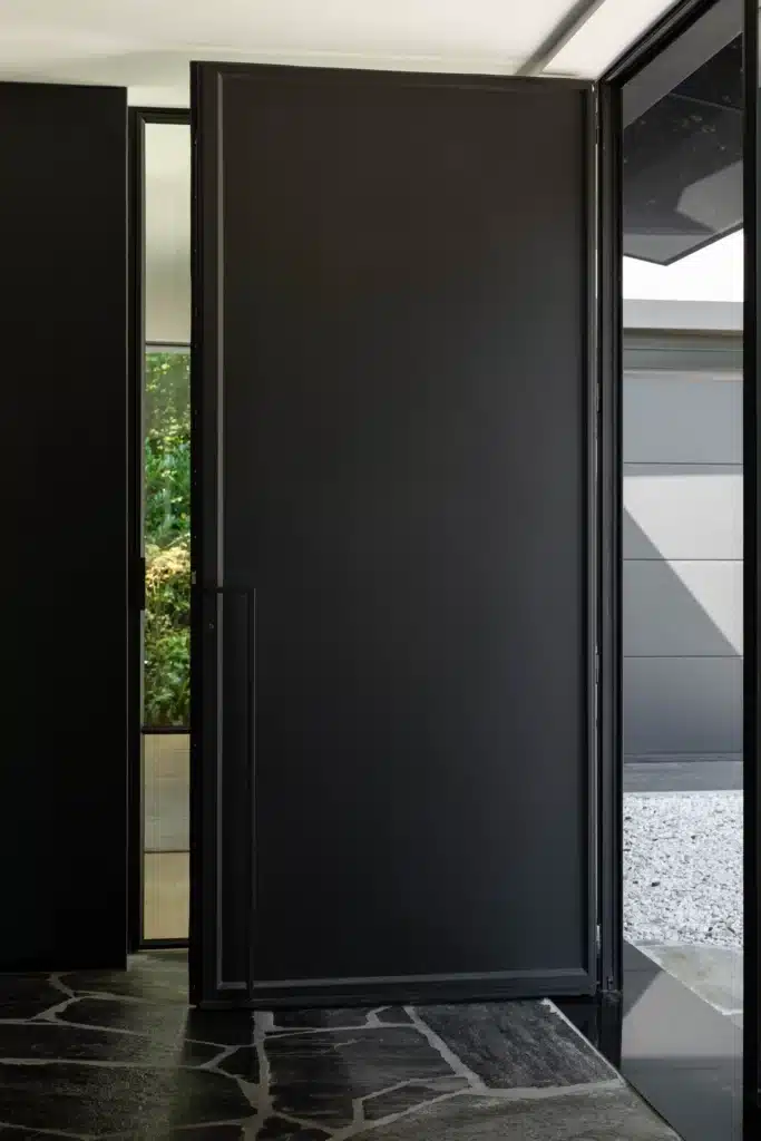 Grote stalen voordeur half geopend boven een terras in natuursteen, met een smal zijlicht ernaast.