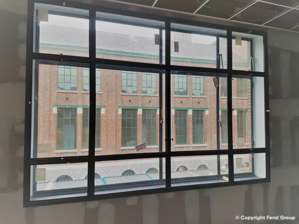 Zwart stalen voorzetraam met roedeverdeling aan de binnenkant van een historisch raam, met zicht op de mijngebouwen.