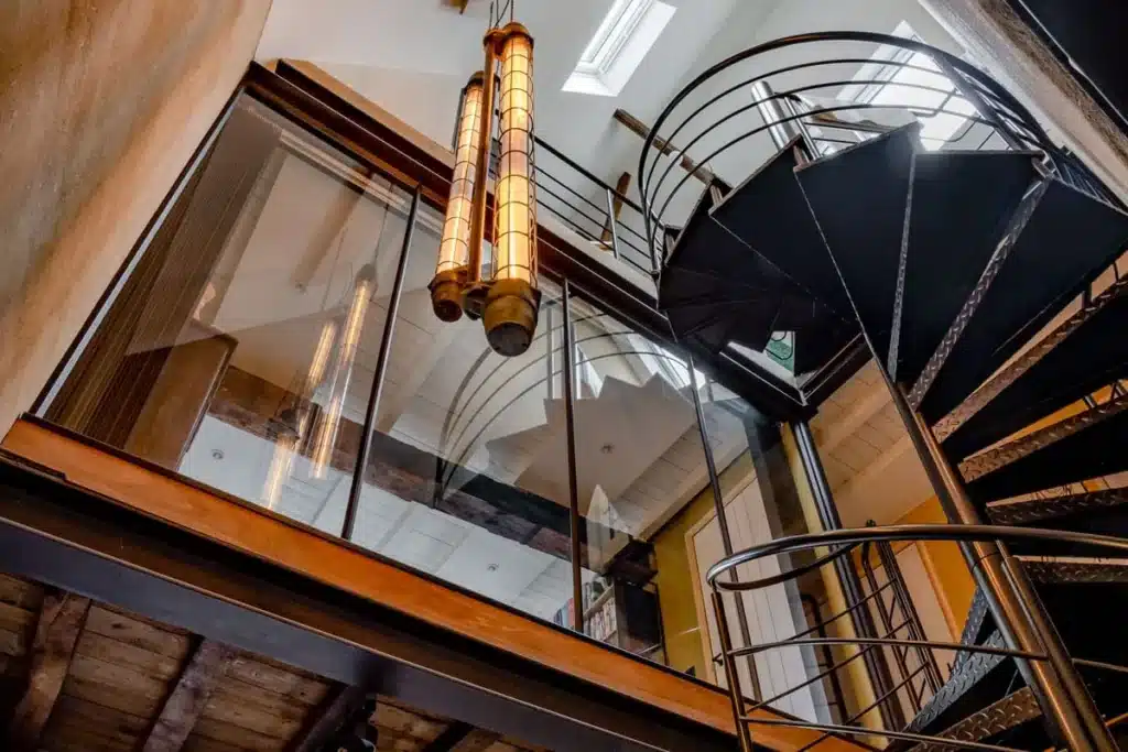 Stalen wenteltrap in een hoge traphal met glazen wanden rond de vides, onder een oud balkenplafond.