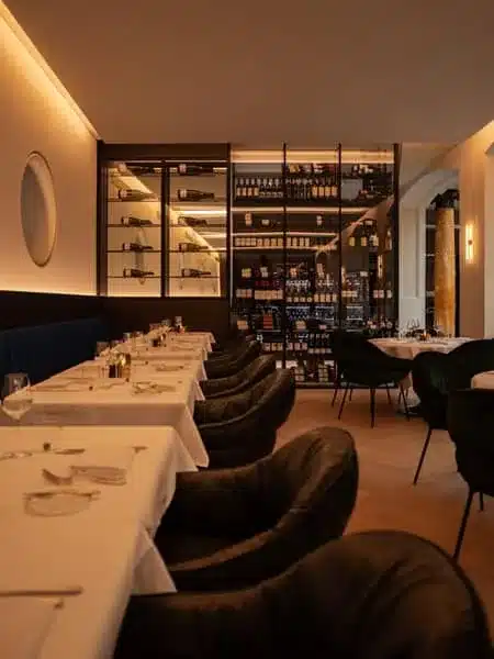 Glazen wijnkamer in zwart staal aan het einde van de restaurantzaal, met verlichte flessenrekken.