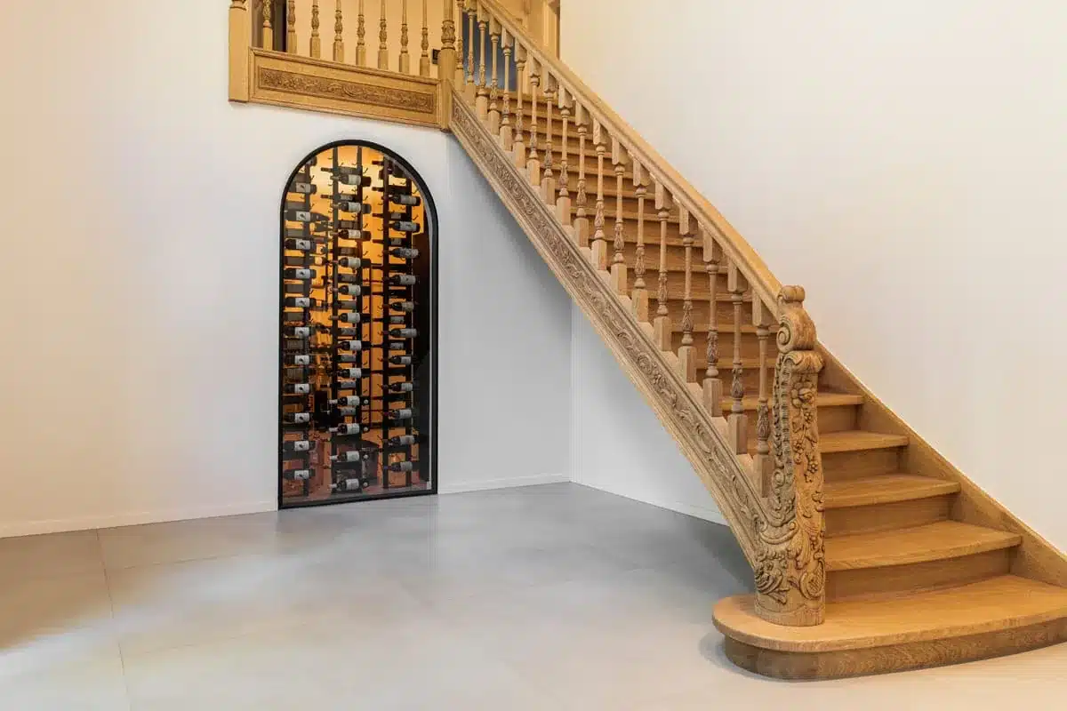 Stalen wijnkast met rondboog en glas onder een houten bordestrap in de inkomhal van een villa.