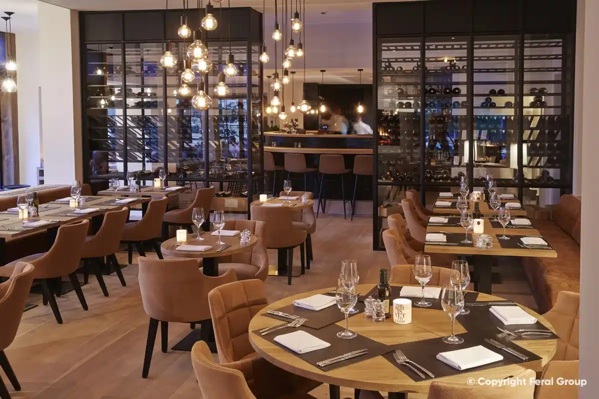 Stalen wijnkasten op maat als visuele afscheiding tussen de bar en de eetzaal, onder een cluster hanglampen.