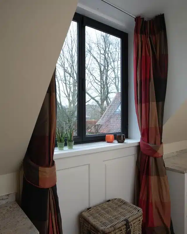 Thermisch onderbroken stalen raam in een dakkapel, met zicht op de bomen en de pannendaken van de hoeve.