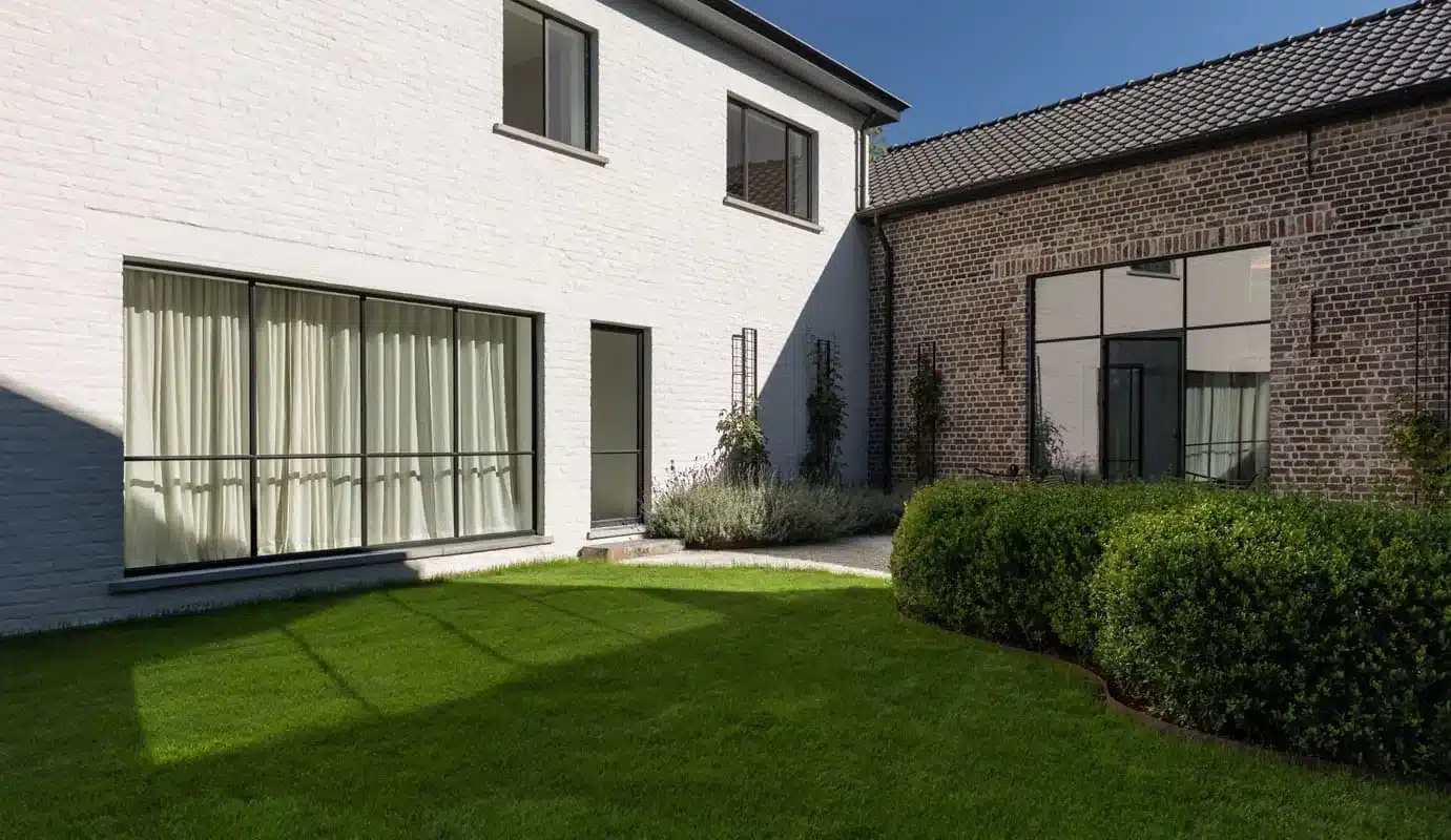 Witte gevel met brede stalen ramen en linnen gordijnen, met gazon en buxushagen voor de showroom.