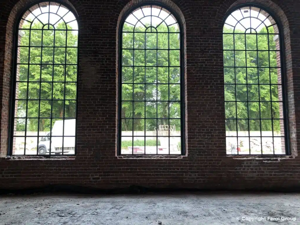 Binnenzicht op drie XL boogramen met fijne roedeverdeling in de bakstenen muur, met zicht op het groen.
